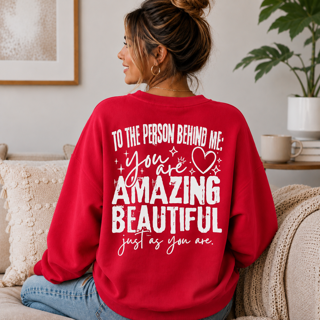 A woman sitting on a couch wearing a red sweatshirt with a white printed quote that reads, 'TO THE PERSON BEHIND ME: you are AMAZING BEAUTIFUL just as you are.'.