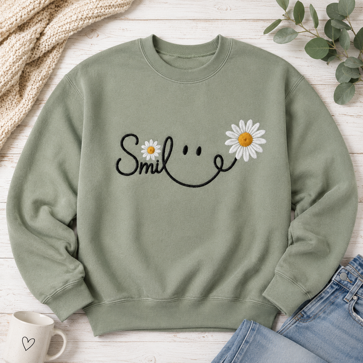 An olive green crewneck sweatshirt with a black smiley face graphic and white daisy flowers, styled in a flat lay on a white wood background with a mug, jeans, and eucalyptus.