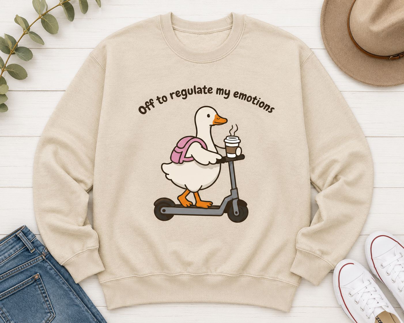 A beige crewneck sweatshirt laid flat on a white wood background, featuring a cartoon duck riding a scooter with the text 'Off to regulate my emotions'. The sweatshirt is styled with blue jeans, white sneakers, a tan hat, and green leaves.