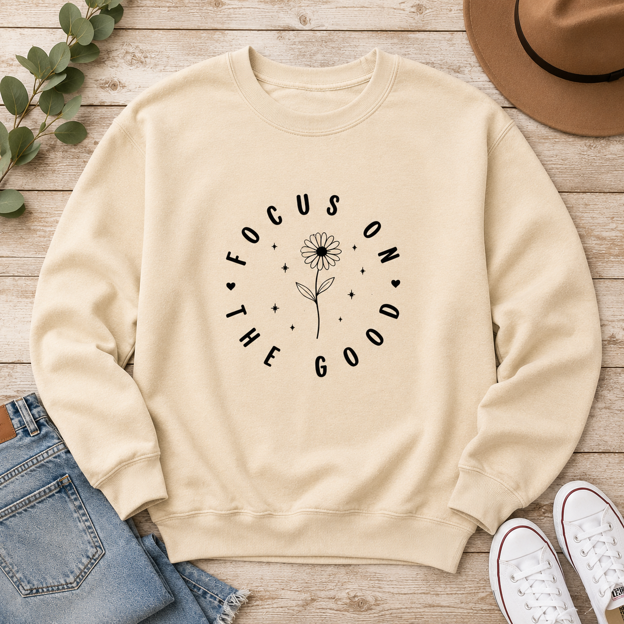 A cream-colored crewneck sweatshirt with a black graphic reading 'FOCUS ON THE GOOD' around a daisy flower, styled in a flat lay on a wooden background with jeans, white sneakers, a hat, and eucalyptus leaves.