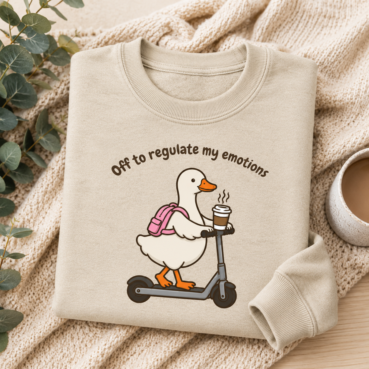 A folded cream sweatshirt with a cartoon duck on a scooter and the text 'Off to regulate my emotions' is styled on a beige knit blanket with eucalyptus leaves and a cup of coffee.
