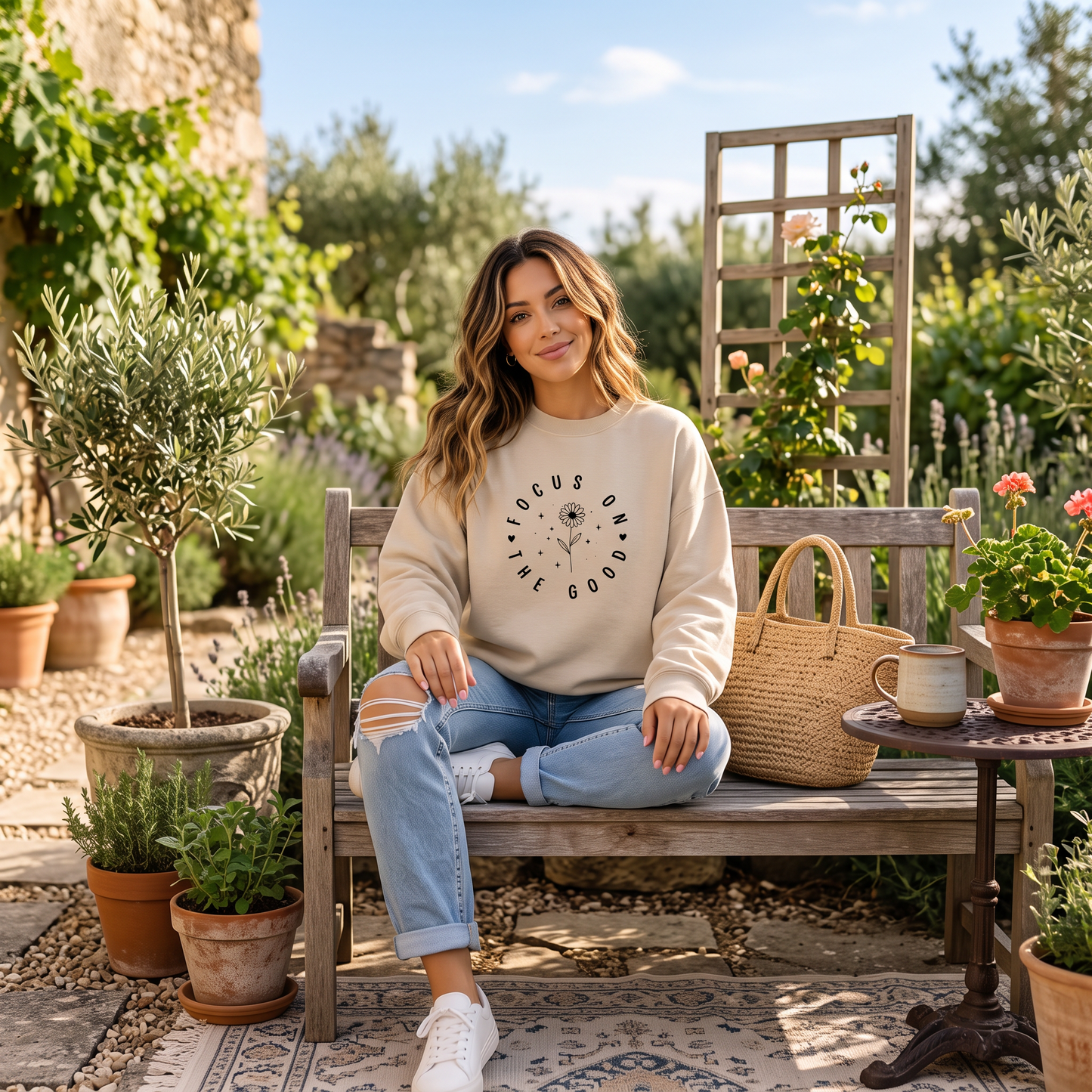 A woman sits on a wooden bench in a garden setting, wearing a cream sweatshirt with a black circular graphic and light blue ripped jeans. A woven straw tote bag sits on the bench next to her.