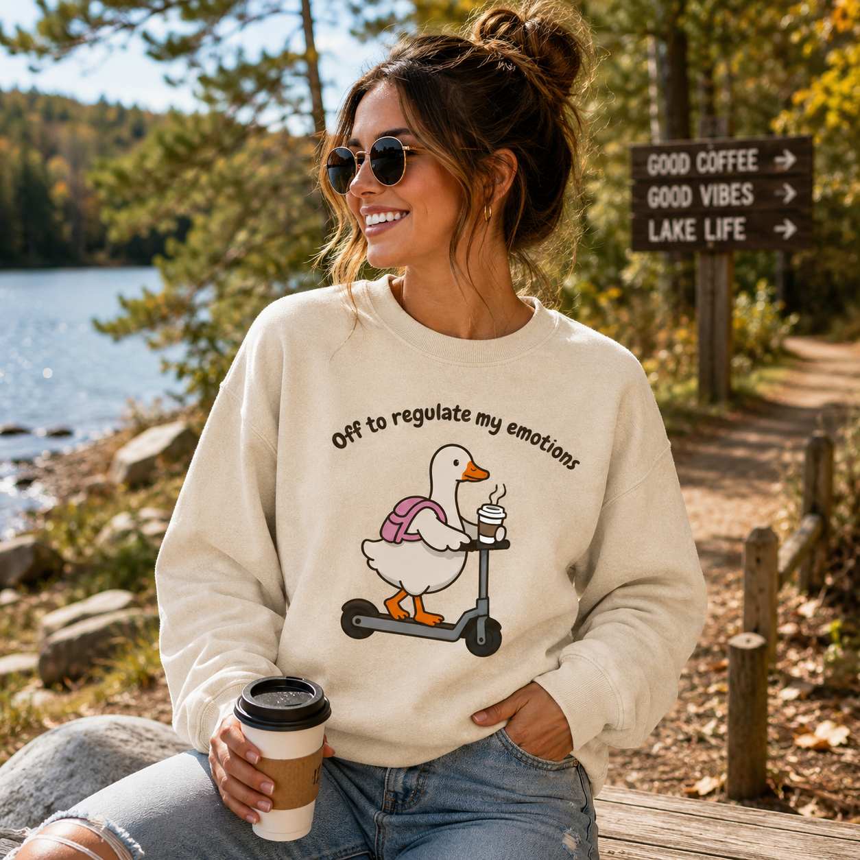A woman wearing a cream-colored sweatshirt with a cartoon duck on a scooter graphic and the text 'Off to regulate my emotions' sits outdoors by a lake holding a coffee cup.