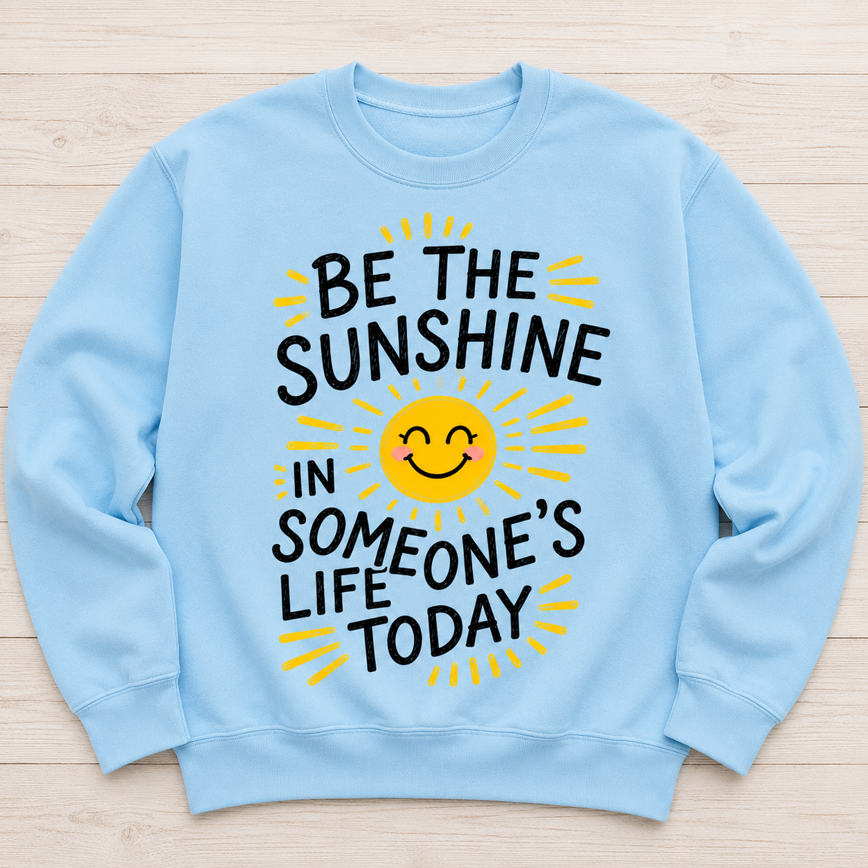A light blue crewneck sweatshirt laid flat on a light wood background, featuring a graphic design with the text 'BE THE SUNSHINE IN SOMEONE'S LIFE TODAY' in black and a smiling yellow sun in the center.