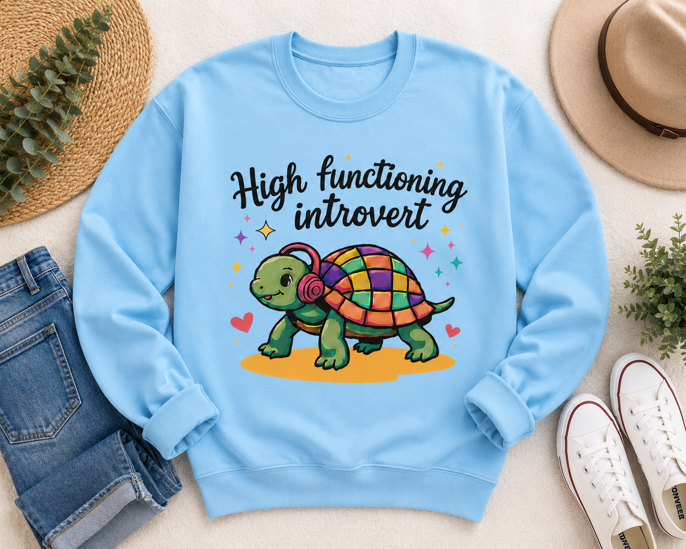 A light blue crewneck sweatshirt laid flat on a beige surface, featuring a colorful cartoon turtle and the text 'High functioning introvert.' The sweatshirt is styled with jeans, white sneakers, a straw hat, and small green plants.