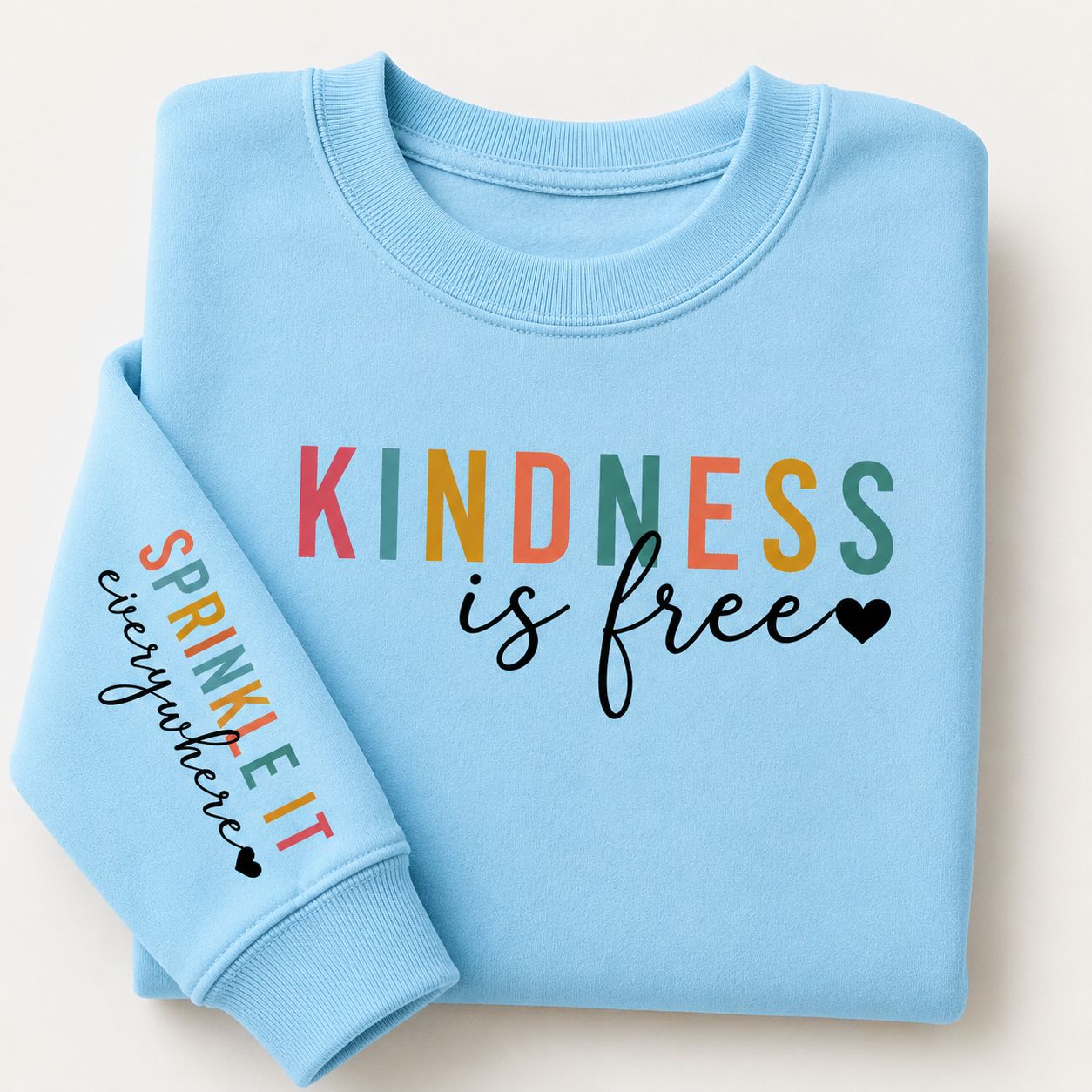 A folded light blue crewneck sweatshirt on a light beige background. The front has the words 'KINDNESS is free' in multicolor and black script, and the left sleeve has 'SPRINKLE IT everywhere' in multicolor and black script. The Scoobie Shak