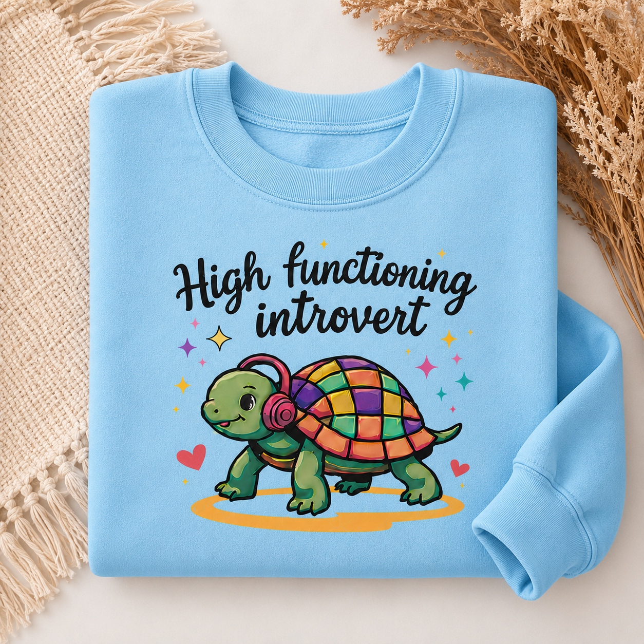 A light blue crewneck sweatshirt laid flat on a light surface with decorative woven textiles and dried grass. The sweatshirt features a colorful cartoon turtle wearing pink headphones with the text 'High functioning introvert' in black script.