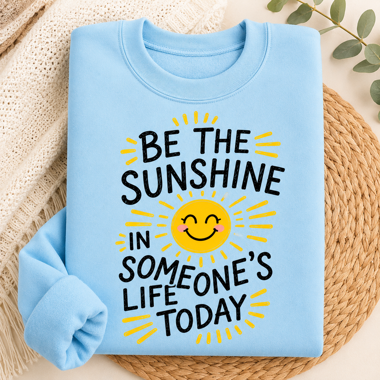 A folded light blue crewneck sweatshirt with a graphic print that reads 'BE THE SUNSHINE IN SOMEONE'S LIFE TODAY' around a smiling yellow sun, styled on a woven placemat with a cream knit blanket.