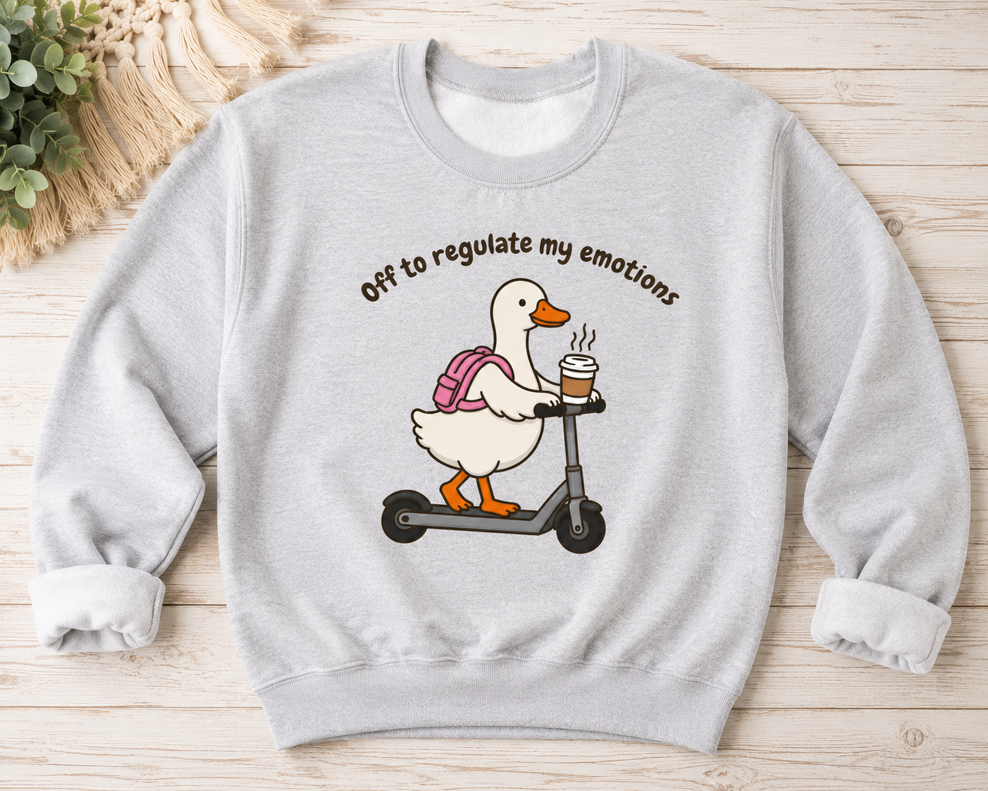 A light gray crewneck sweatshirt laid flat on a light wood background, featuring a cartoon graphic of a white duck riding a scooter with a pink backpack and holding a coffee cup, with the text 'Off to regulate my emotions' above it.