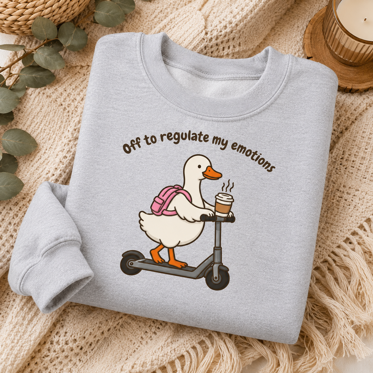 A light heather gray sweatshirt laid flat on a beige knit blanket, featuring a cartoon white duck on a scooter with a pink backpack and the text 'Off to regulate my emotions.'.