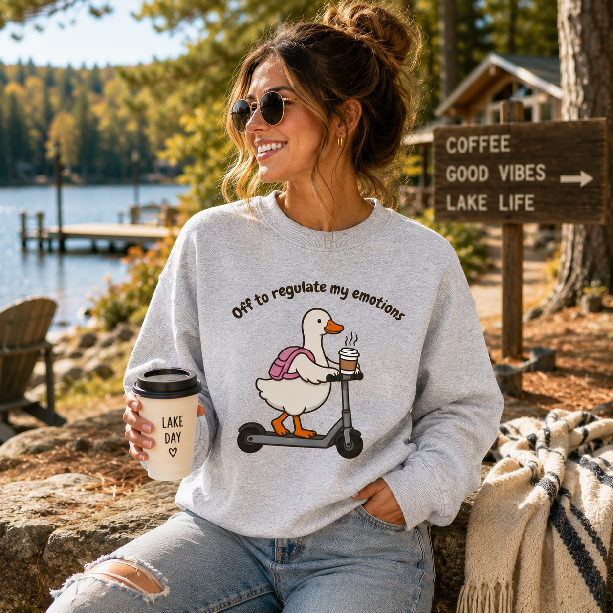 A woman sitting outdoors by a lake wearing a light gray sweatshirt with a duck graphic on a scooter and the text 'Off to regulate my emotions.' She holds a coffee cup and jeans, with a wooden sign reading 'COFFEE GOOD VIBES LAKE LIFE' in the background.