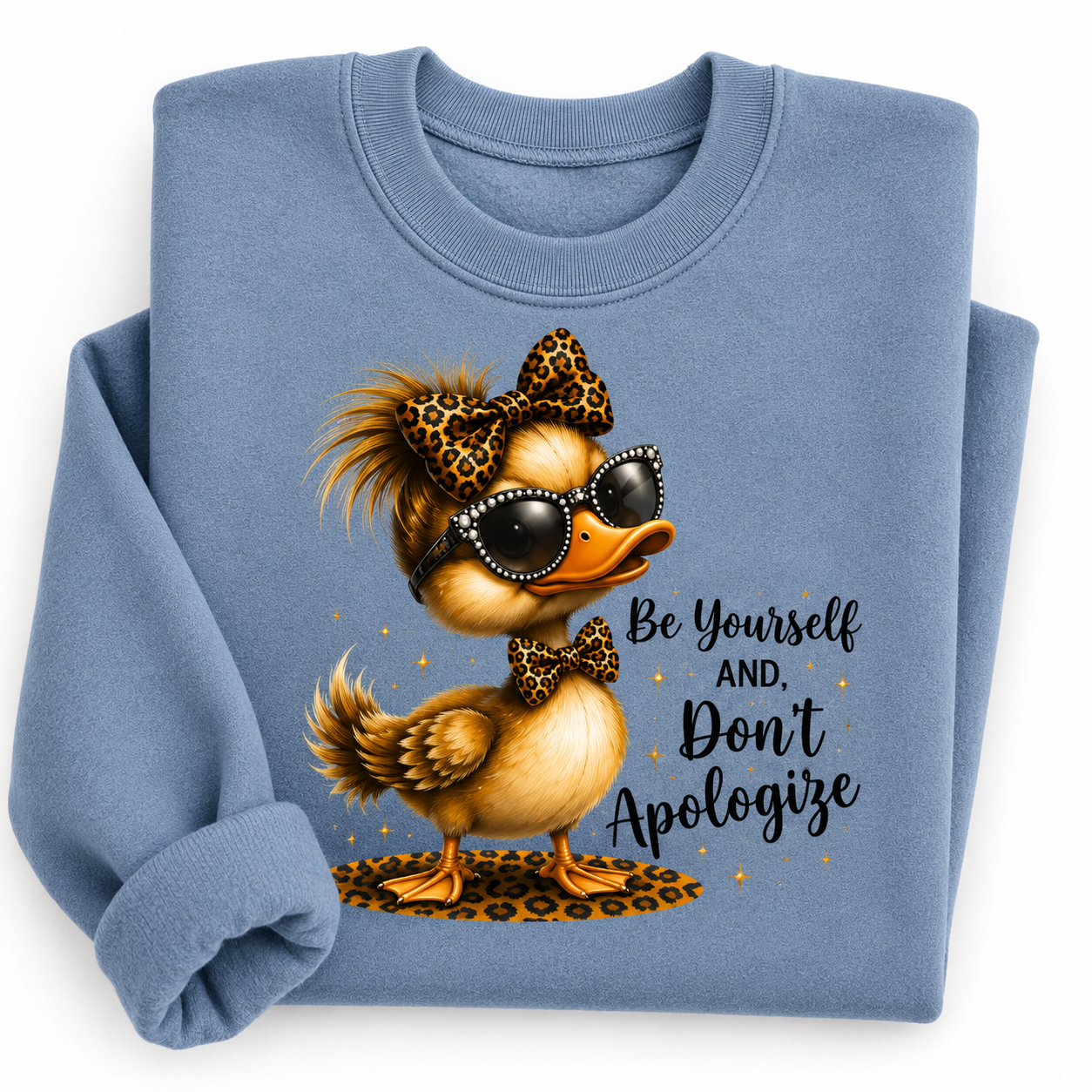 A folded light blue crewneck sweatshirt featuring a graphic of a stylish duck wearing leopard-print sunglasses, sunglasses, and bow tie, with the text 'Be Yourself AND, Don't Apologize' printed on the front.