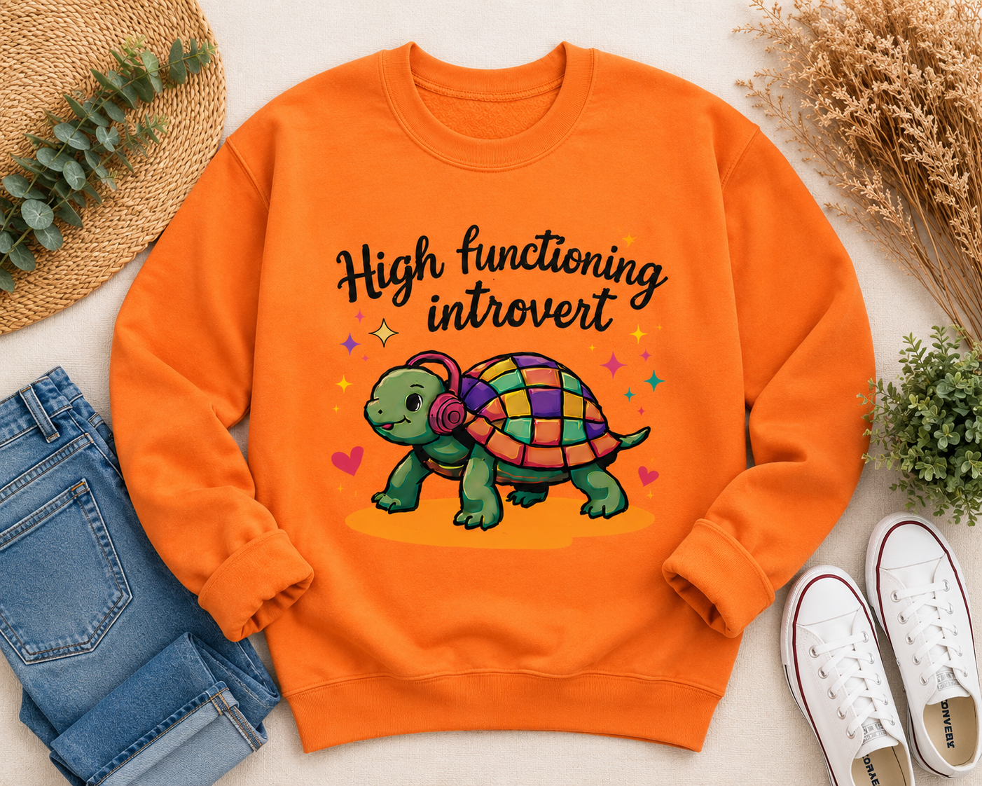 An orange crewneck sweatshirt with a colorful graphic of a turtle and the text 'High functioning introvert' in black script. The sweatshirt is styled in a flat lay with blue jeans, white sneakers, a straw hat, and dried flowers on a light beige background.