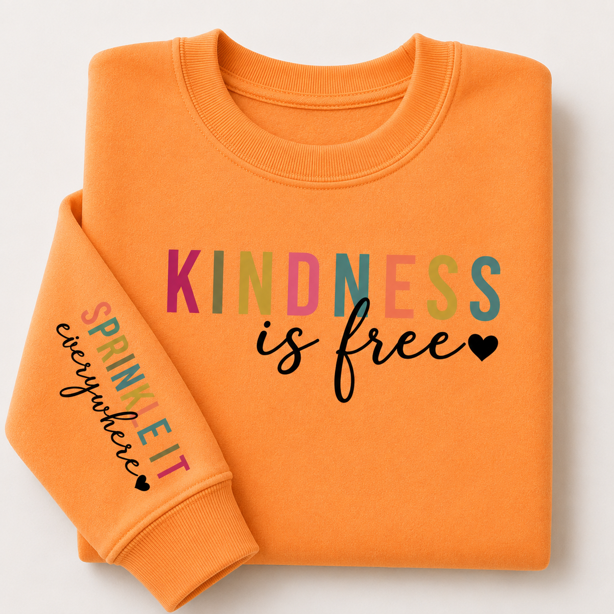 An orange crewneck sweatshirt with a folded sleeve, featuring the text 'KINDNESS is free' in multicolor and black script on the front, and 'SPRINKLE IT everywhere' in multicolor and black script on the sleeve.