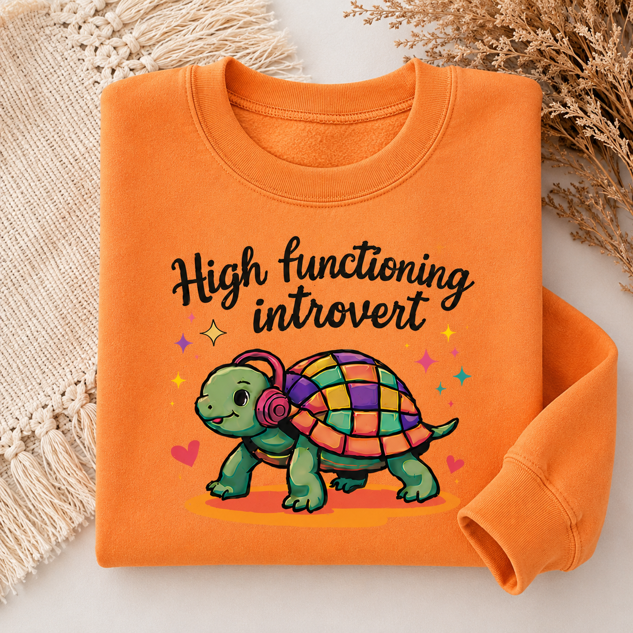 An orange crewneck sweatshirt laid flat on a light surface, styled with a beige woven textile and dried flowers. The sweatshirt features a graphic of a green turtle with a colorful checkered shell and headphones, with the text 'High functioning introvert' in black script.