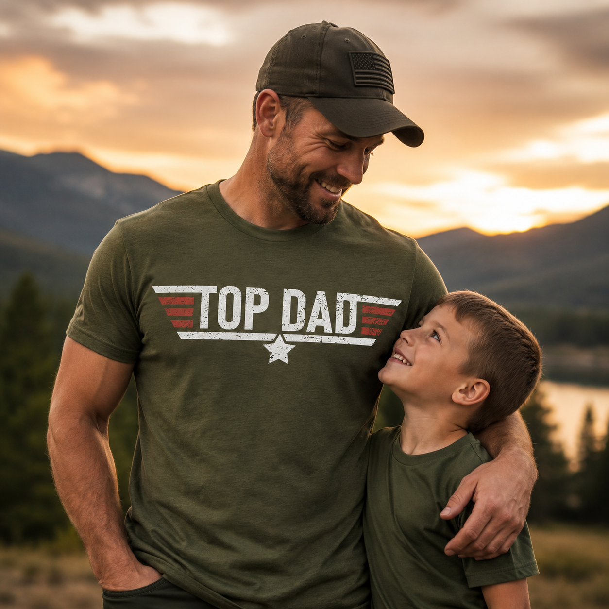 Man wearing a 'Top Dad' t-shirt with a child in front of a mountainous landscape during sunset. The Scoobie Shack