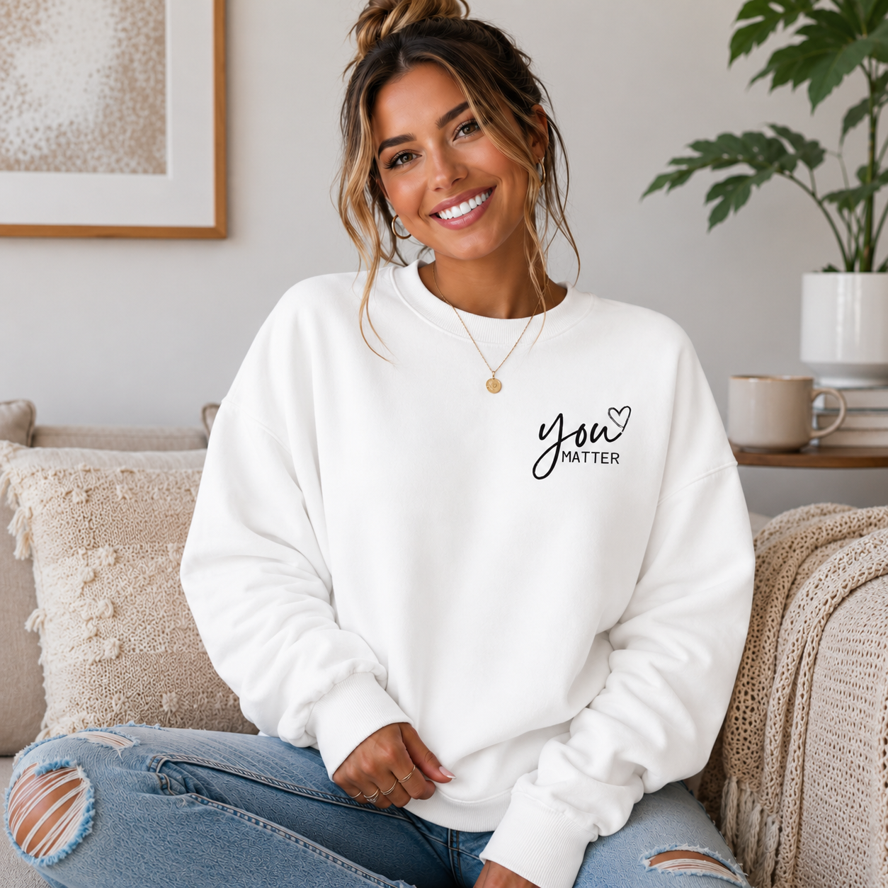 A woman sitting on a beige couch wearing a white crewneck sweatshirt with the black text 'you MATTER' printed on the left chest, paired with distressed blue jeans.