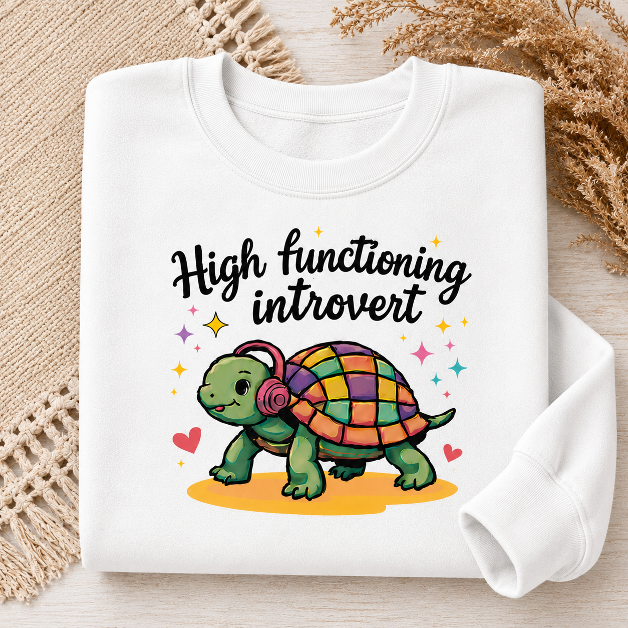 A folded white long-sleeve sweatshirt on a light wood surface with decorative woven mats and dried grass. The sweatshirt features a colorful graphic of a green turtle with a multicolored checkered shell, pink headphones, and the text 'High functioning introvert' in black script.