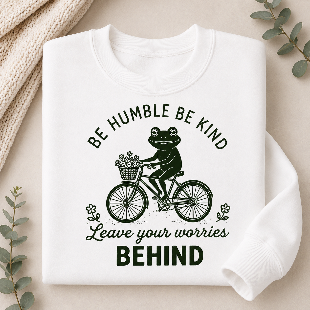 A folded white crewneck sweatshirt with a black graphic print of a frog riding a bicycle with flowers, accompanied by the text 'BE HUMBLE BE KIND Leave your worries BEHIND'. The sweatshirt is styled in a flat-lay arrangement with a beige knit blanket, eucalyptus leaves, and a neutral background.