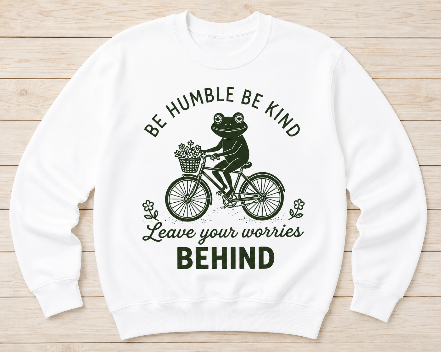 A white crewneck sweatshirt with a dark green graphic of a frog riding a bicycle and the text 'BE HUMBLE BE KIND Leave your worries BEHIND', displayed flat on a light wood plank background.