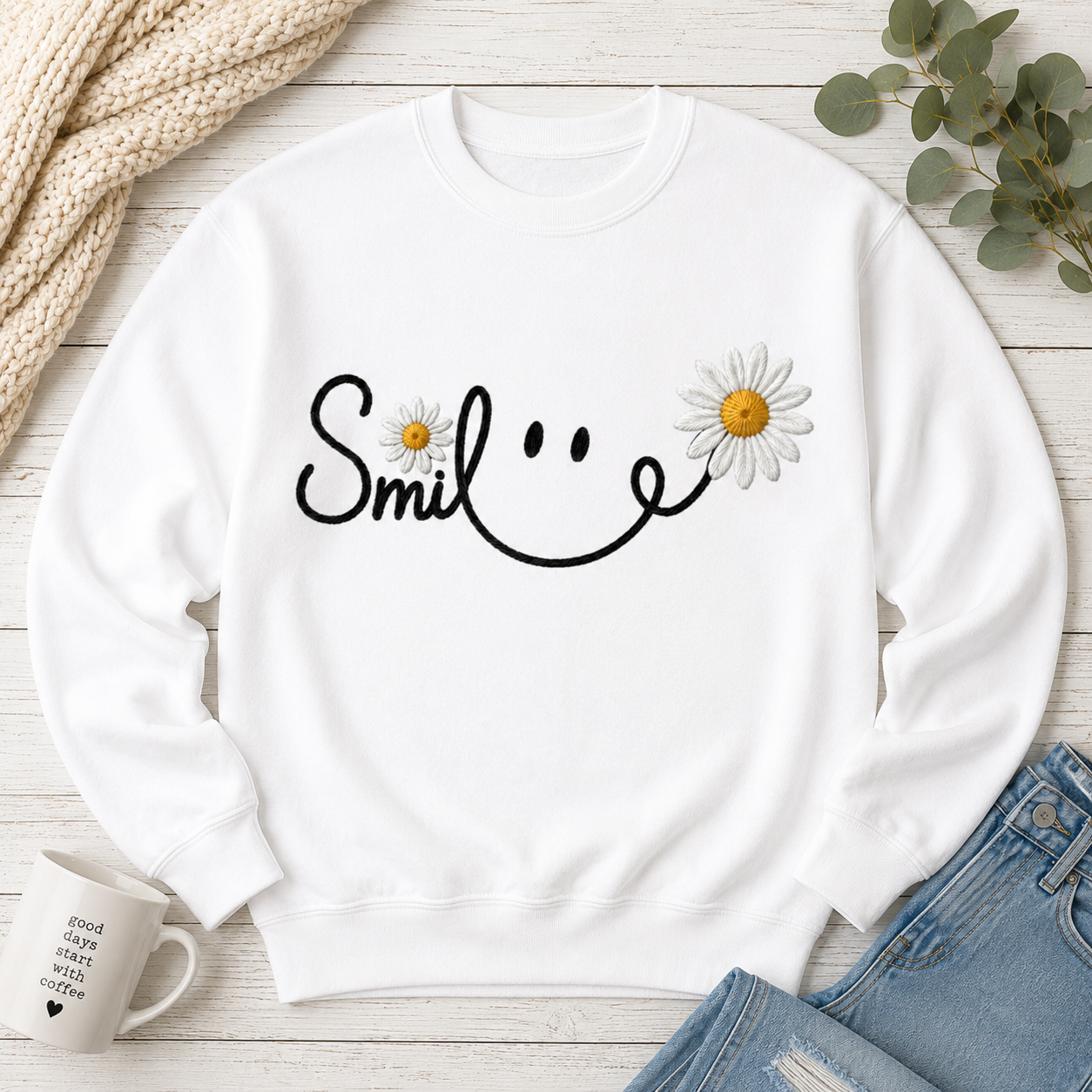 A white crewneck sweatshirt laid flat on a light wood surface, featuring a black smiley face graphic with two daisy flowers. The sweatshirt is styled with a beige knit blanket, green eucalyptus leaves, a white mug, and blue jeans.