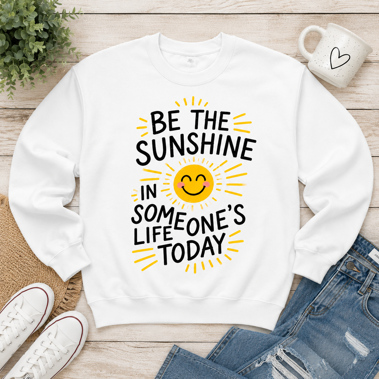 A white crewneck sweatshirt with a yellow and black graphic reading 'BE THE SUNSHINE IN SOMEONE'S LIFE TODAY' around a smiling sun face. The sweatshirt is displayed in a flat-lay arrangement on a light wood background with blue jeans, white sneakers, a mug, and a plant.