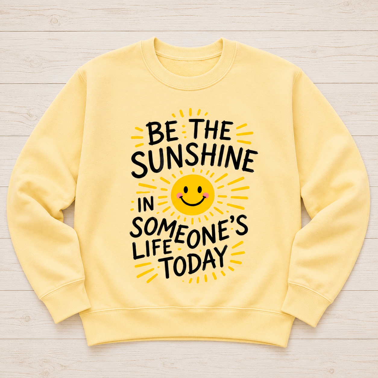 A pale yellow crewneck sweatshirt laid flat on a light wood background, featuring a graphic print that reads 'BE THE SUNSHINE IN SOMEONE'S LIFE TODAY' in black text around a yellow smiling sun.