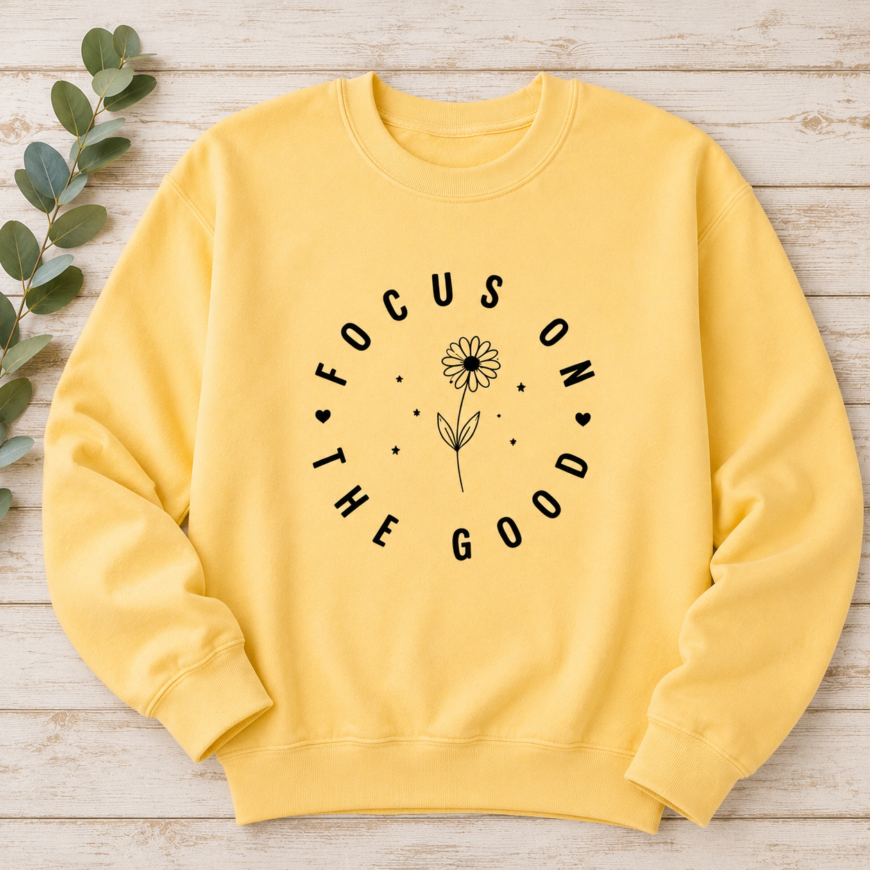 A yellow crewneck sweatshirt with a black graphic print reading 'FOCUS ON THE GOOD' around a flower, laid flat on a light wood background with green leaves on the side.