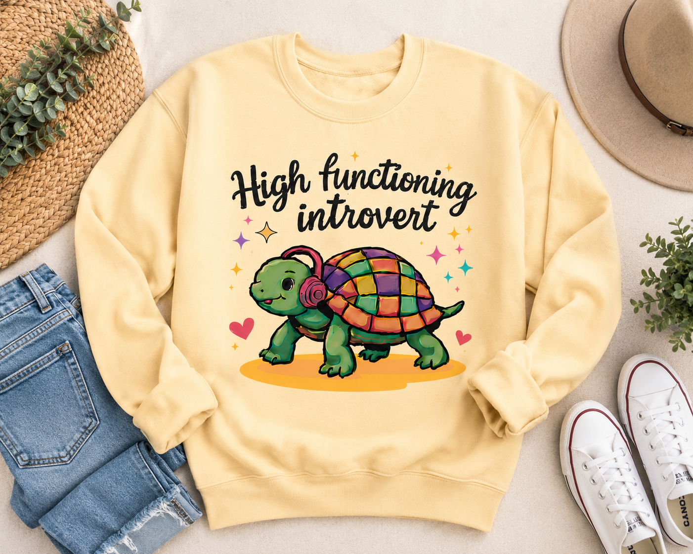 A pale yellow crewneck sweatshirt laid flat on a light beige surface, surrounded by styled props including blue jeans, white sneakers, a straw hat, and small green plants. The sweatshirt has a colorful graphic of a turtle wearing over-ear headphones with the text 'High functioning introvert' above it.