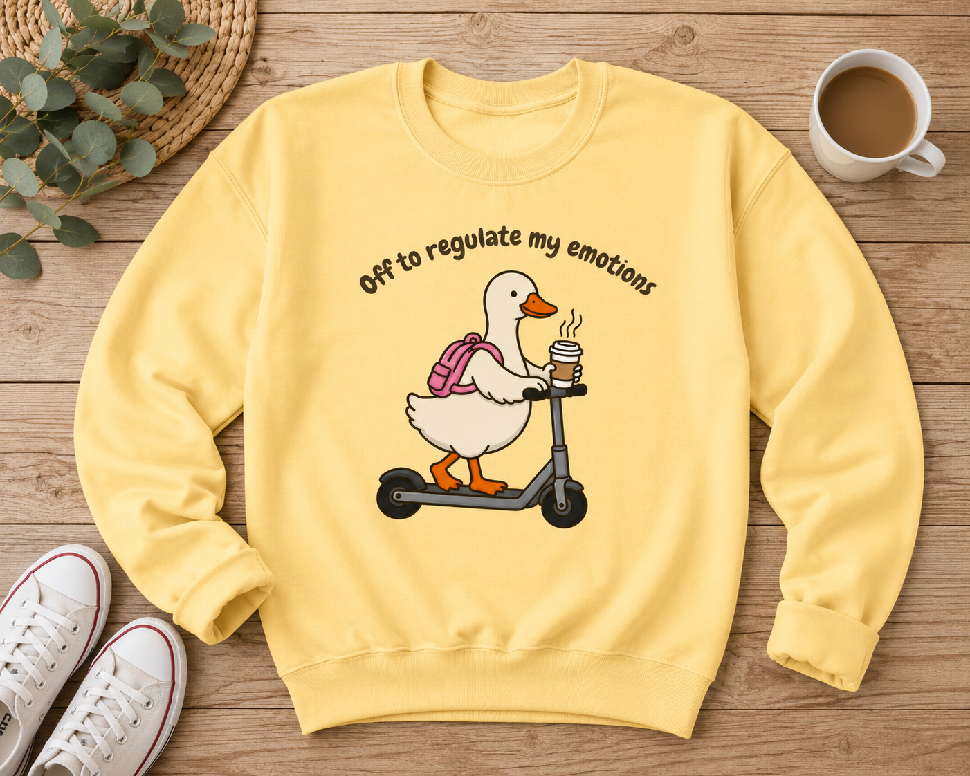 A pale yellow crewneck sweatshirt laid flat on a wooden surface, featuring a cartoon duck riding a scooter with the text 'Off to regulate my emotions' above it. The sweatshirt is styled with a cup of coffee, white sneakers, a woven mat, and greenery.
