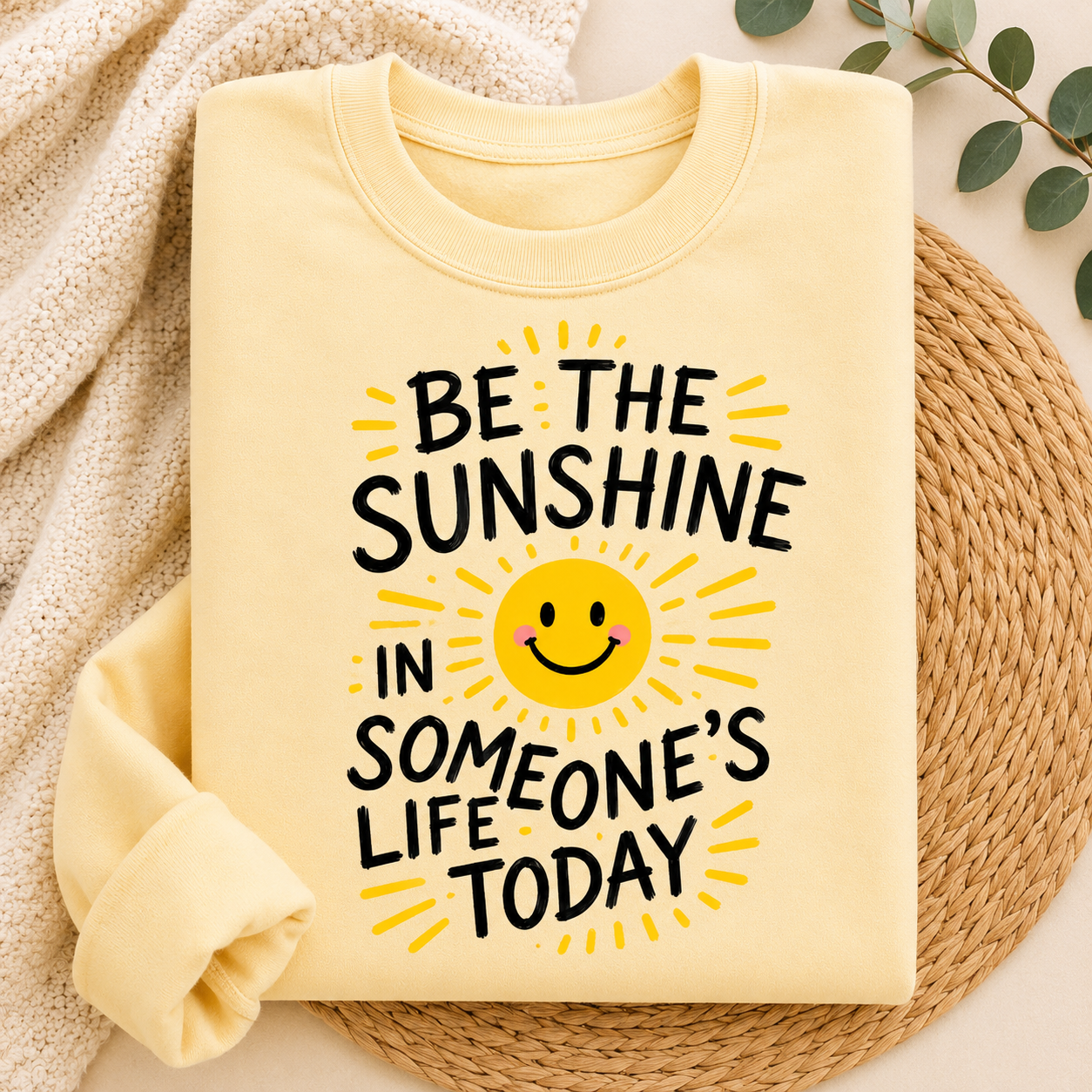 A folded pale yellow crewneck sweatshirt with a graphic that reads 'BE THE SUNSHINE IN SOMEONE'S LIFE TODAY' around a smiling sun face. The sweatshirt is styled on a woven round mat with a cream knit blanket and green leaves in the background.