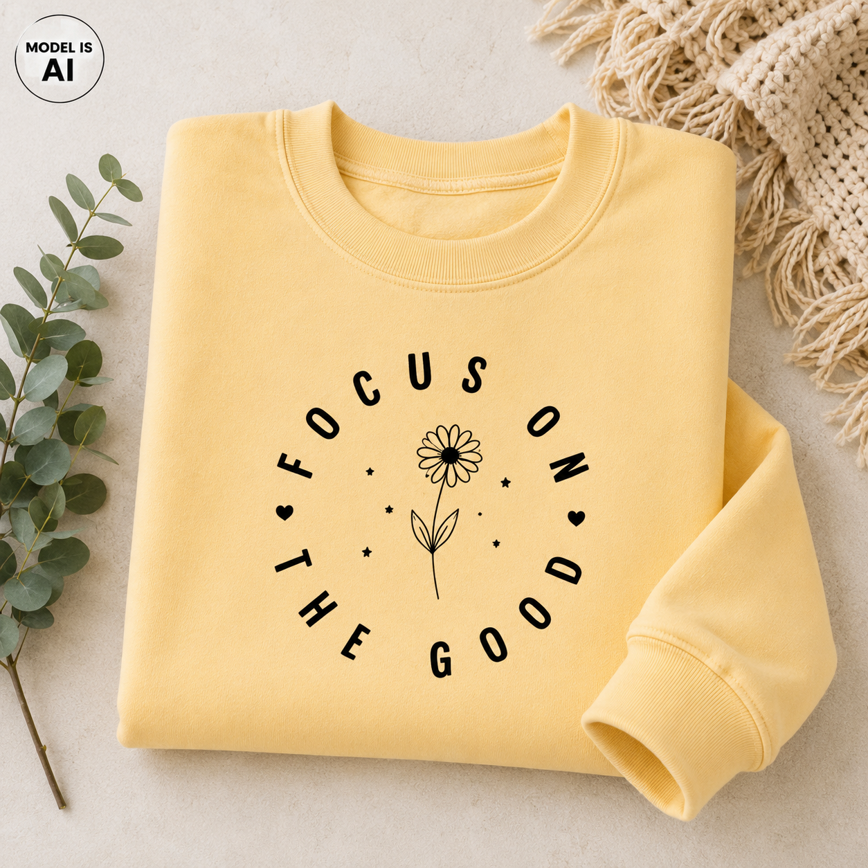 A folded pale yellow sweatshirt with a black graphic reading 'FOCUS ON THE GOOD' around a flower illustration, styled on a light beige surface with eucalyptus leaves and a knitted textile.