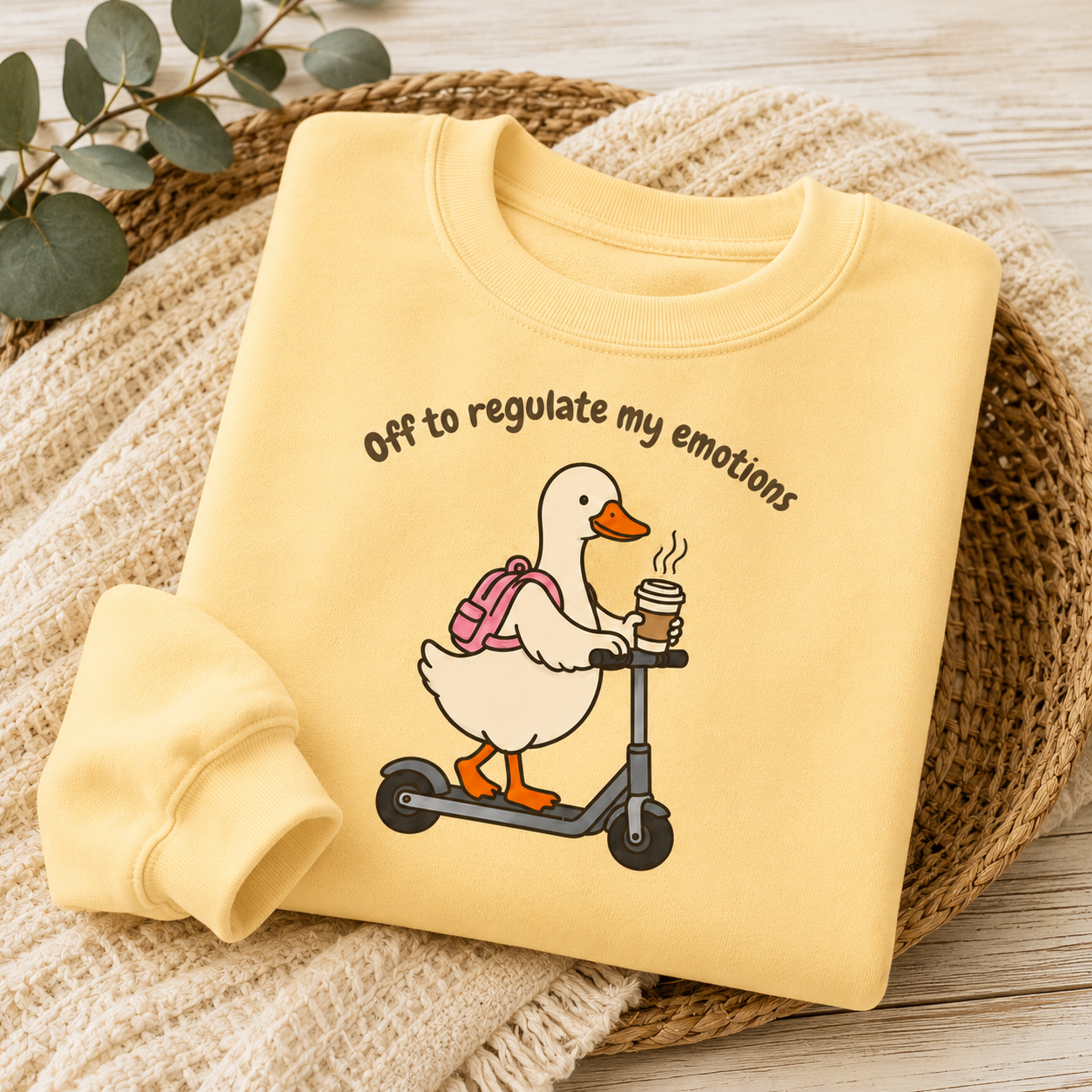 A folded pale yellow sweatshirt with a cartoon duck riding a scooter and holding a coffee cup, with the text 'Off to regulate my emotions' printed above. The sweatshirt is styled on a woven mat with a knit blanket and eucalyptus leaves.