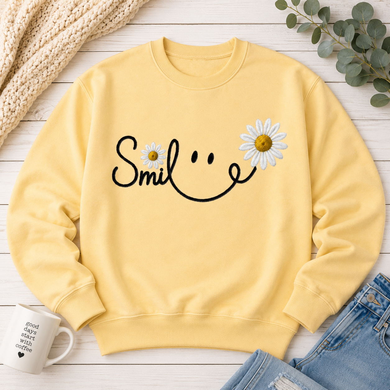 A pale yellow crewneck sweatshirt laid flat on a white wood surface, featuring a black smiley face graphic made from the word 'Smile' and two white daisy flowers with yellow centers. Styled flat-lay props include a chunky cream knit blanket, a white mug, blue jeans, and eucalyptus leaves.