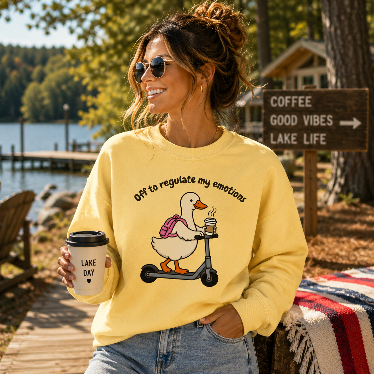 A woman wearing a pale yellow sweatshirt with a duck graphic on a scooter and the text 'Off to regulate my emotions' stands outdoors by a lake holding a coffee cup. The background includes trees, a wooden sign, and a picnic blanket.